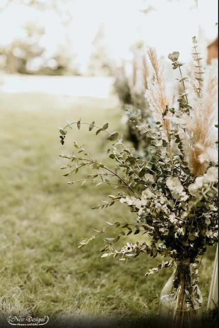 A wedding Aisle, in a field, Lined with Tall Glass Milk Jug Style Vases, overflowing with Baby Eucalyptus Greens and Tall Nude Pampas Grass Stems